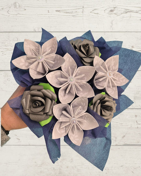 A bouquet of handmade origami paper flowers, featuring lavender five-petal blossoms and gray paper roses, wrapped in blue tissue paper and held by a hand against a white wood background.