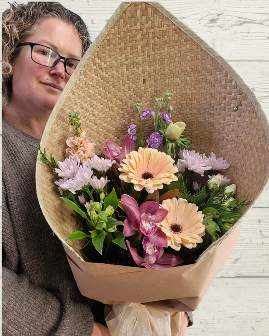 A person holding a large, beautifully arranged flower bouquet wrapped in a natural woven paper cone. The bouquet features a mix of pastel pink, purple, peach, and green flowers, set against a light wood paneled background.