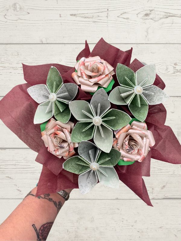 A bouquet of artificial flowers wrapped in dark burgundy tissue paper and held by a tattooed arm against a white wood plank background. The bouquet features light pink roses and five silver-gray paper hydrangeas with white centers.