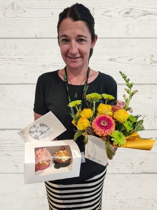 person holding a floral arrangement, cupcakes and a handmade card.