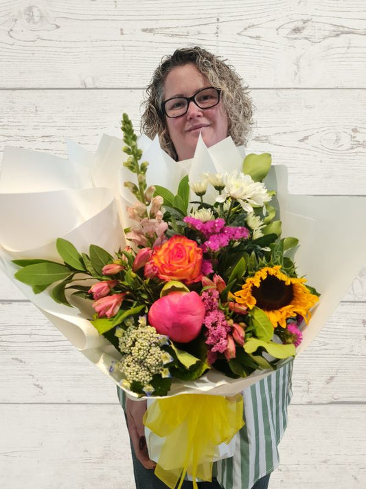 A bouquet of assorted fresh flowers including pink roses, sunflowers, and other seasonal flowers, arranged in a white paper vase with a yellow bow.