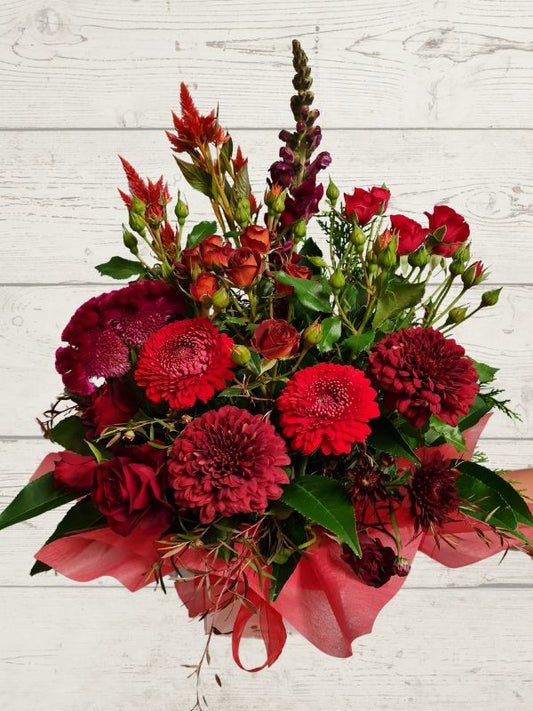 A red-themed floral arrangement featuring various red flowers and green foliage, arranged in a container with red wrap.