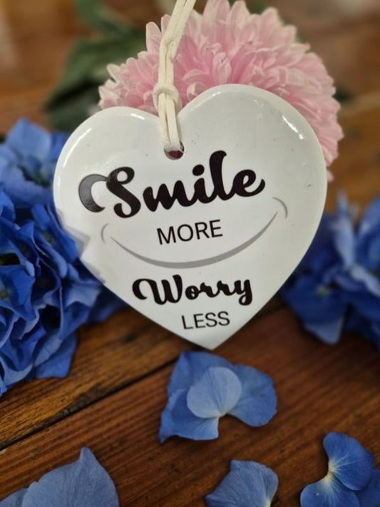 A ceramic heart-shaped plaque with the words 'SMILE MORE WORRY LESS' written on it, attached to a pink flower.