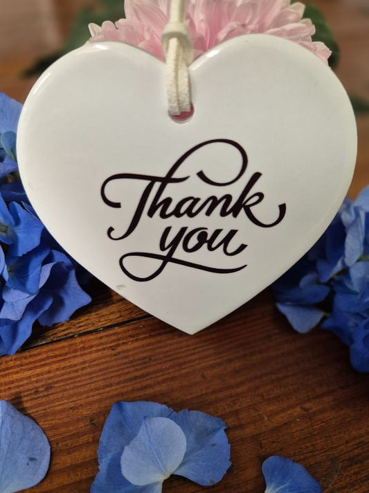 A white ceramic heart-shaped plaque with the words 'Thank you' written in black script, attached to a pink ribbon, displayed on a wooden surface with blue flower petals scattered around.