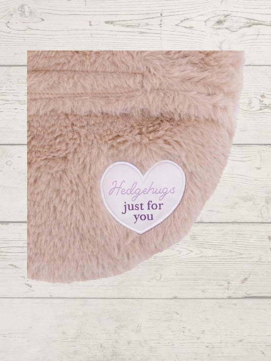 Fluffy pink hedgehog with a heart-shaped card reading 'Hedgehugs just for you' on a wooden surface.
