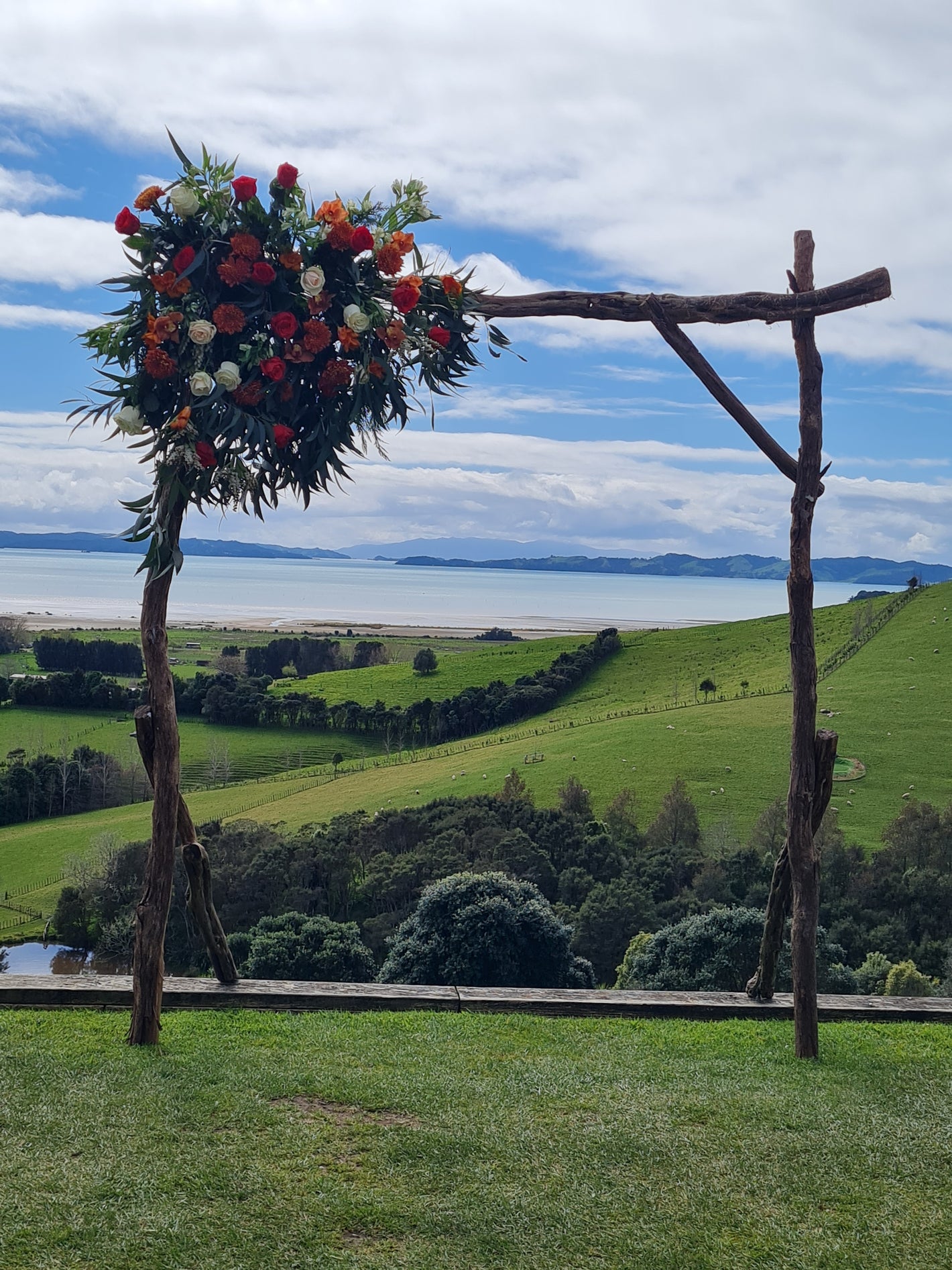 Floral arrangement on a wooden arch against a scenic background with fields and trees.
