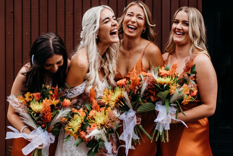 Four women in orange dresses holding bouquets of flowers, laughing together.
