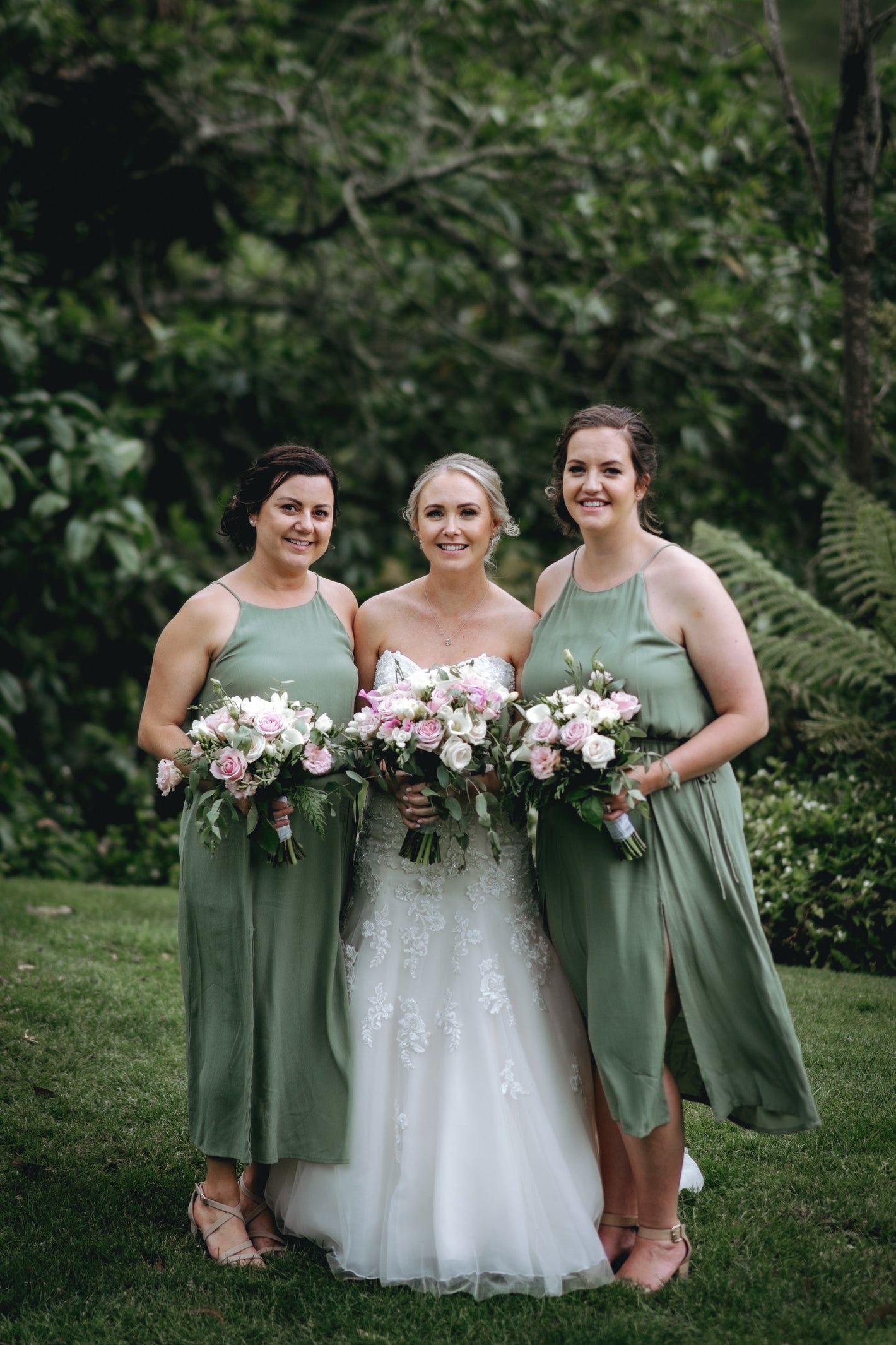 Bride with two bridesmaids in green dresses holding floral bouquets outdoors.