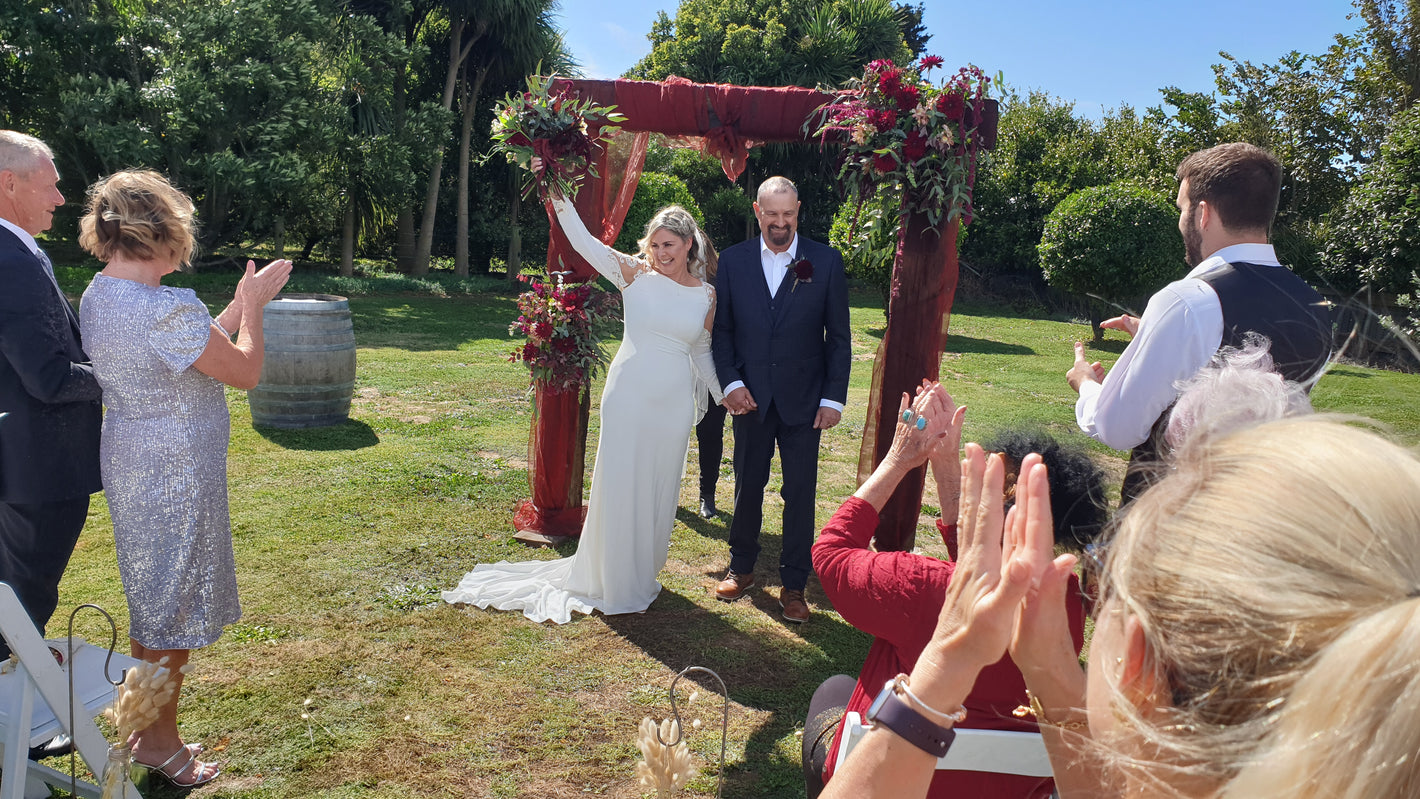 Wedding ceremony outdoors with a couple under a decorated arch, surrounded by guests.