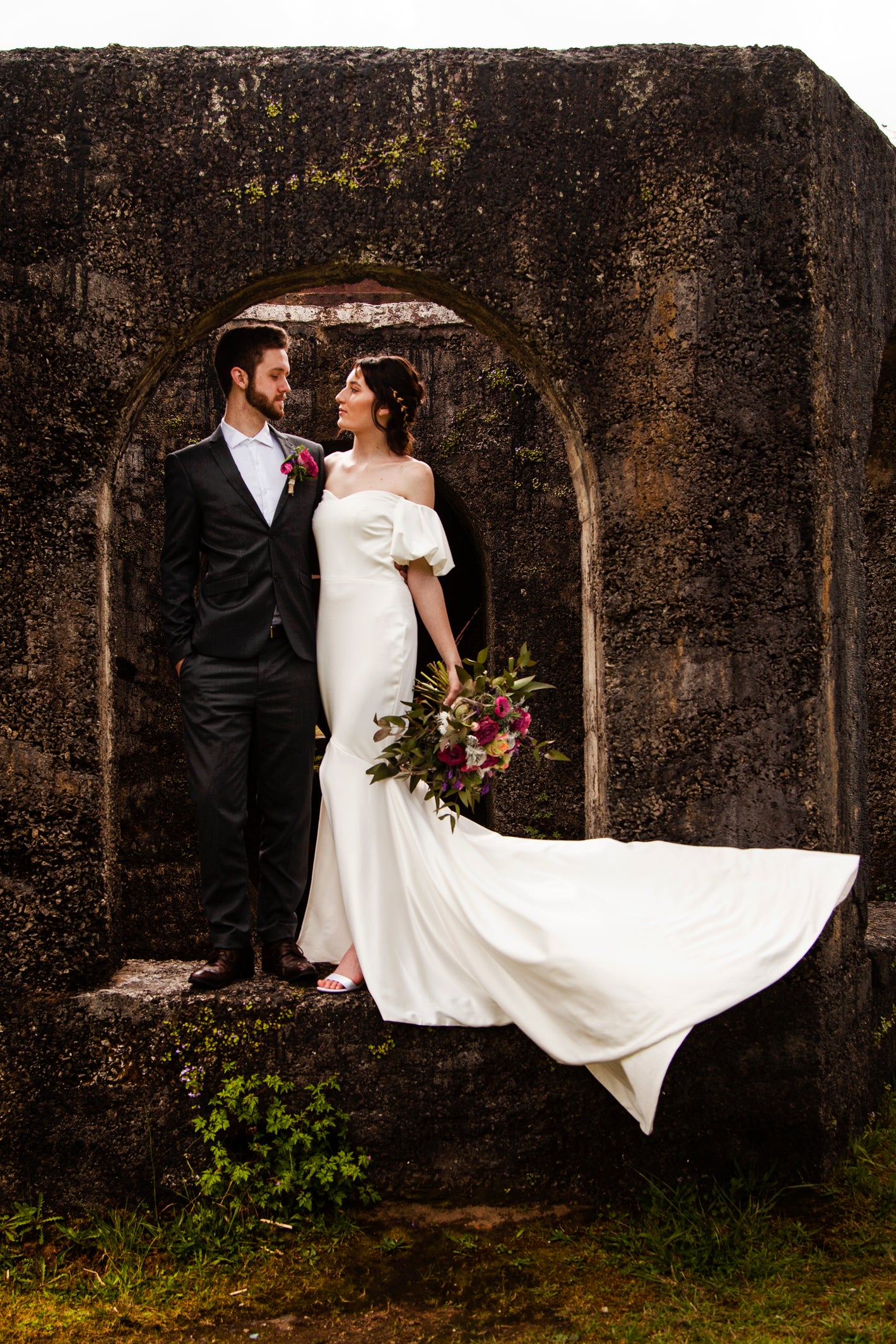 Couple in wedding attire standing in a rustic stone archway, bride holding flowers.
