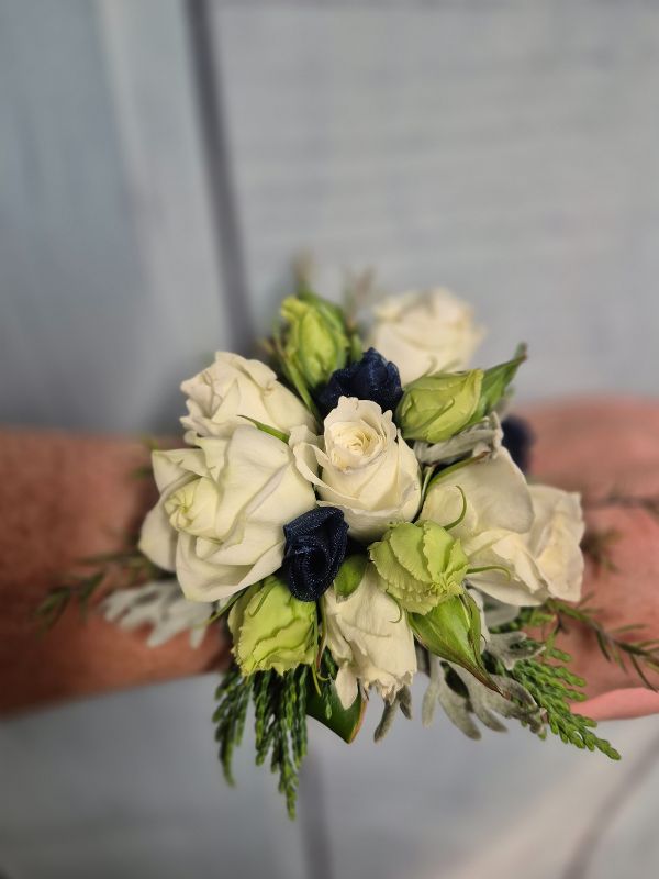 A wrist corsage featuring white and navy flowers with green leaves, attached to a navy organza ribbon.
