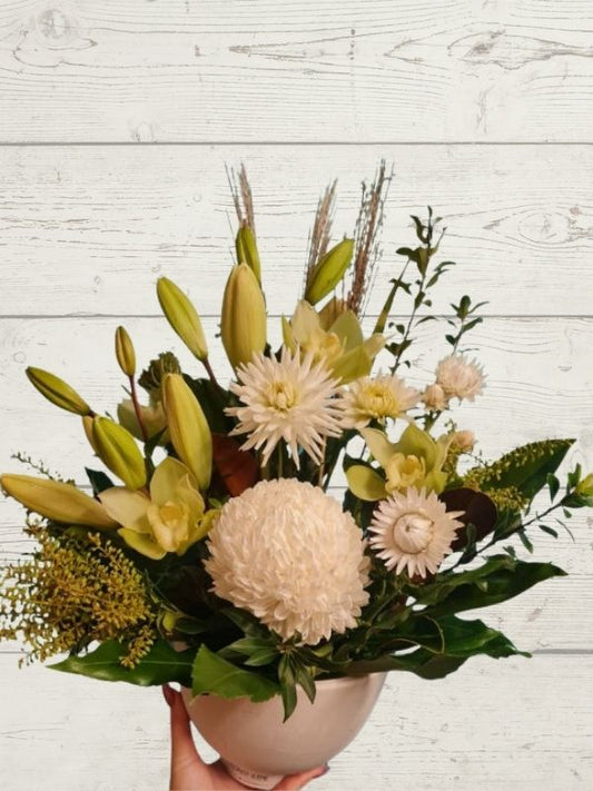 A floral arrangement featuring a variety of fresh flowers including lilies and orchids, in shades of white, green, and beige, presented in a container.