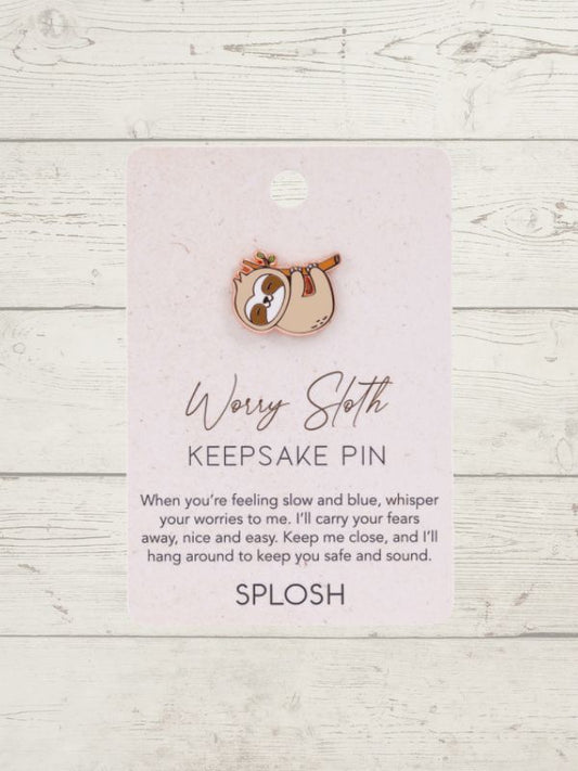 A small enamel keepsake pin shaped like a sloth resting on a pink card that reads 'Worry Sloth KEEPSAKE PIN' with a quote and the brand name 'SPLOSH', photographed against a whitewashed wood background.