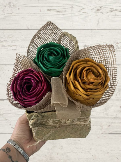 Bouquet of three flax flowers in green, purple, and gold held by a hand on a wooden surface.