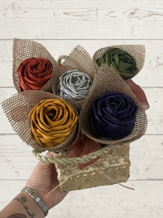 Bouquet of multicolored flax flowers in a woven basket held by a hand on a light wooden surface.