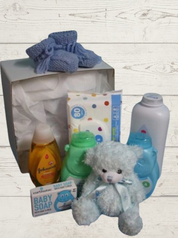 A blue baby gift box opened to display a variety of baby care products including lotions, body wash, shampoo, wipes, a soft toy, and handmade booties.