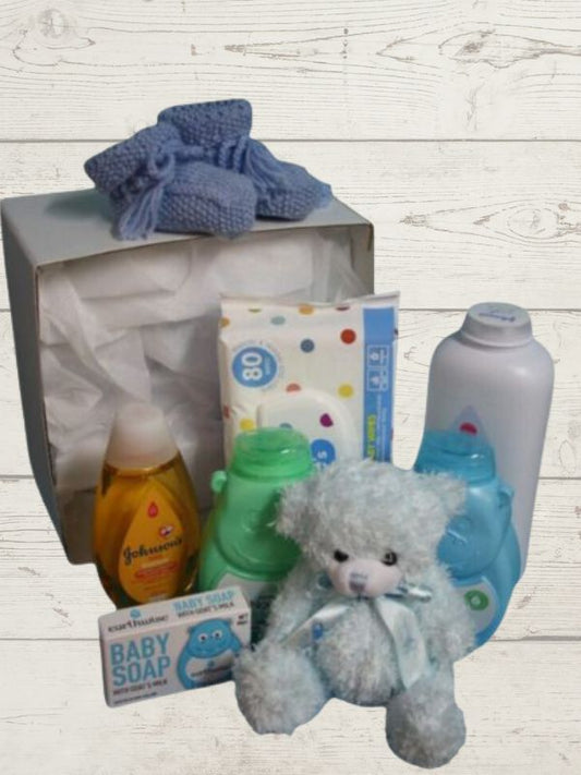 A blue baby gift box opened to display a variety of baby care products including lotions, body wash, shampoo, wipes, a soft toy, and handmade booties.