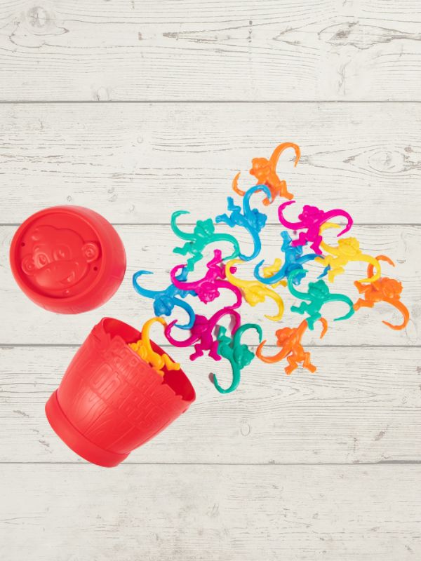 A red Barrel of Monkeys game with colorful plastic monkeys spilled out onto a wooden surface.