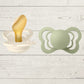 Two BIBS pacifiers with an anatomical glow design, one pacifier with a green ring on the shield and the other with a black ring, placed on a textured surface.