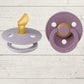 Two BIBS brand pacifiers, one in purple and one in blue, each with a round shield and a ring handle. The purple pacifier has a gold-colored nipple, and both display the BIBS logo.