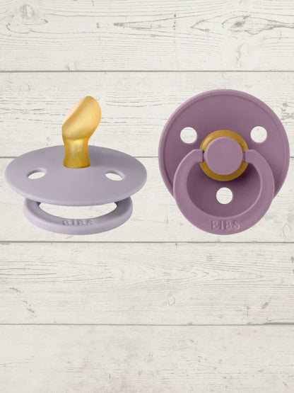 Two BIBS brand pacifiers, one in purple and one in blue, each with a round shield and a ring handle. The purple pacifier has a gold-colored nipple, and both display the BIBS logo.