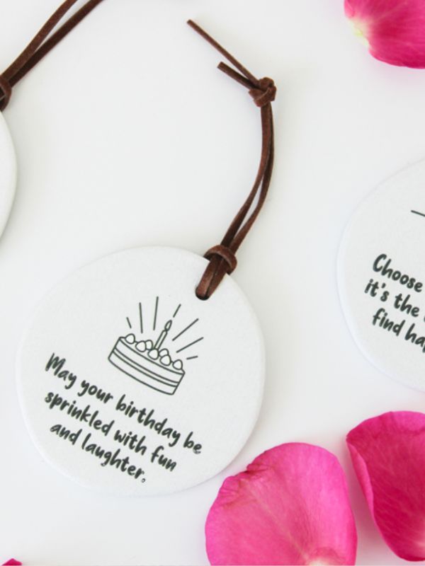A round, white ceramic plaque with embossed text and a graphic of candles, rope for hanging, placed on a surface with pink rose petals around it.
