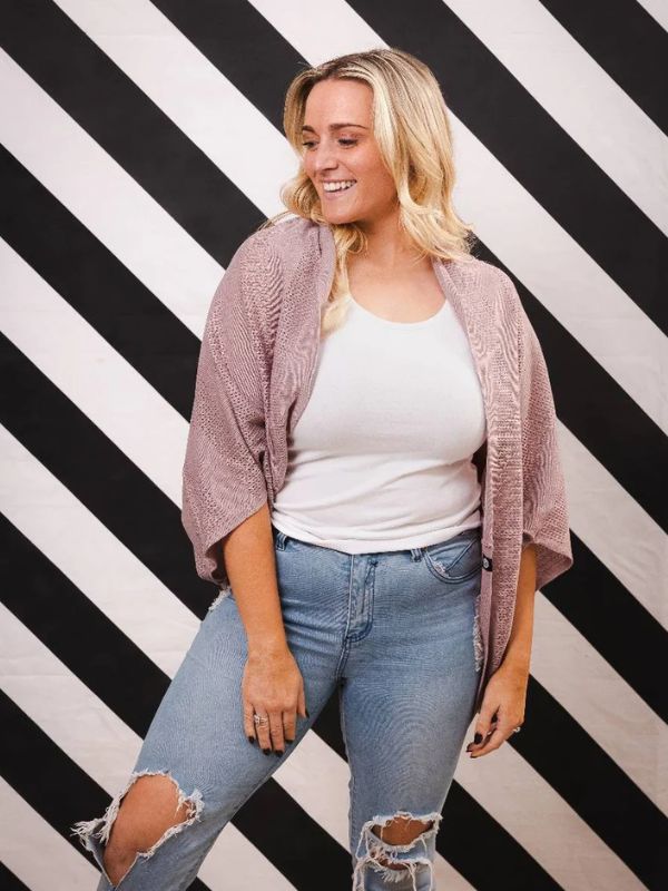 A woman standing against a black and white striped background, wearing a pink cardigan over a white top and blue jeans with a ripped design.