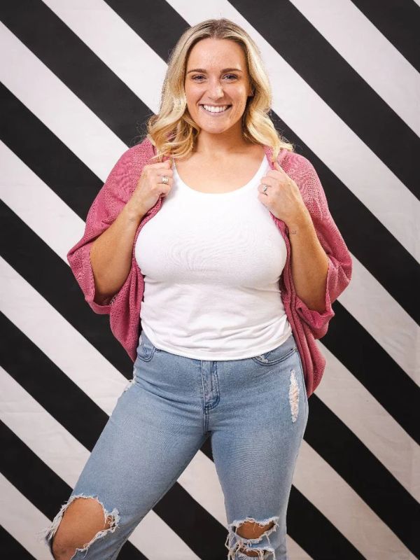 A woman standing against a black and white striped background, wearing a pink cardigan over a white top and blue jeans with a ripped design.