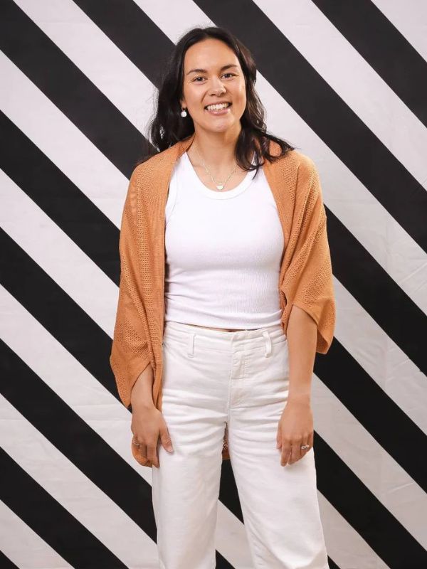 A woman standing against a black and white striped background, wearing a brown cardigan over a white top and white jeans.