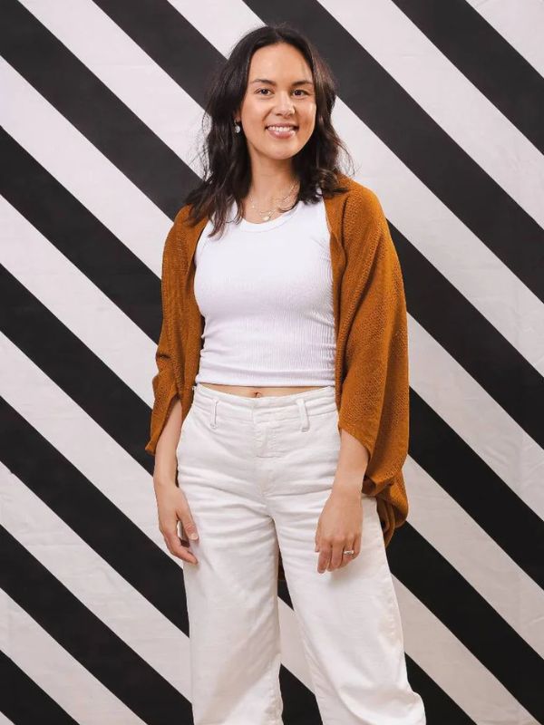A woman standing against a black and white striped background, wearing a dark brown cardigan over a white top and white jeans with a ripped design.