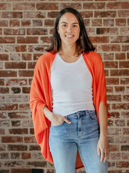 A person standing in front of a brick wall, wearing a red cardigan over a white top and blue jeans.