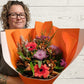 A person holding a hand-tied bouquet of hot pink and orange flowers, wrapped in orange paper.
