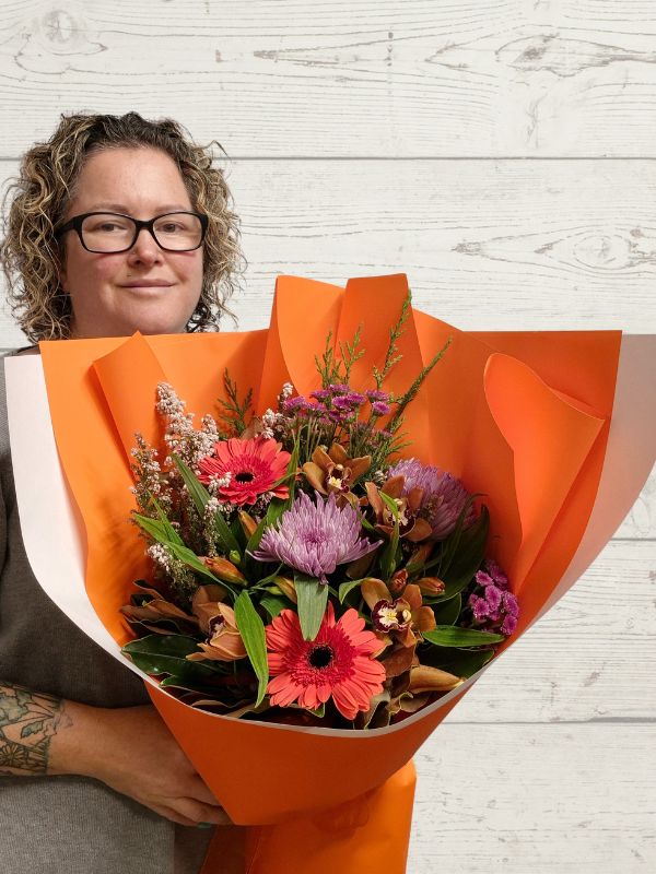 A person holding a hand-tied bouquet of hot pink and orange flowers, wrapped in orange paper.