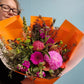 Person holding a bouquet of hot pink and orange flowers wrapped in orange paper against a light blue background