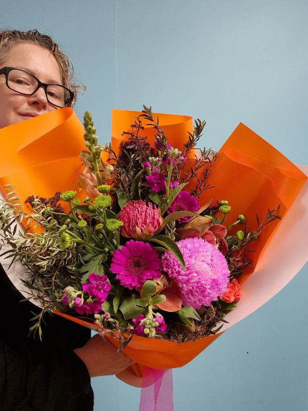 Person holding a bouquet of hot pink and orange flowers wrapped in orange paper against a light blue background