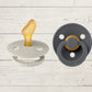 Two BIBS pacifiers with an anatomical glow design, one pacifier with a green ring on the shield and the other with a black ring, placed on a textured surface.