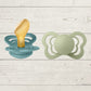 Two BIBS brand pacifiers, one with a yellow nipple and one with a green nipple, placed on a wooden surface.