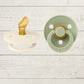 Two BIBS pacifiers with an anatomical glow design, one pacifier with a green ring on the shield and the other with a black ring, placed on a textured surface.