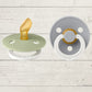 Two BIBS pacifiers with an anatomical glow design, one pacifier with a green ring on the shield and the other with a black ring, placed on a textured surface.