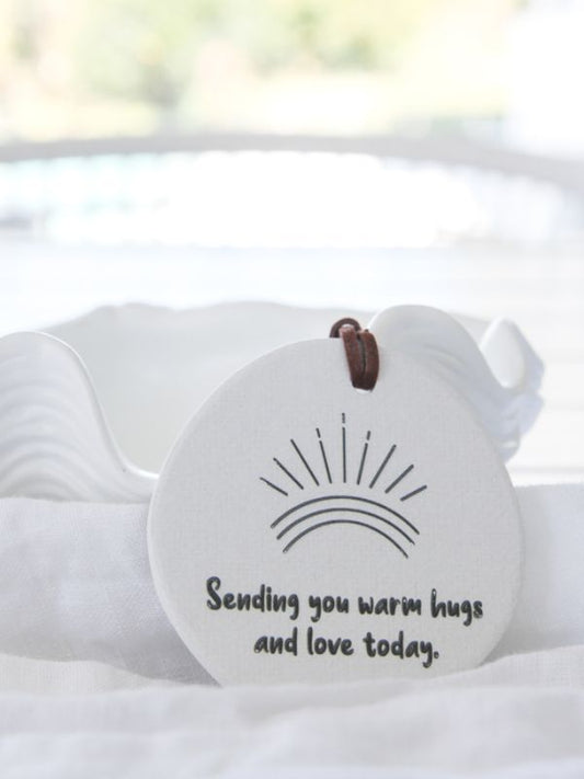A round, white ceramic plaque with embossed text 'SENDING YOU WARM HUGS AND LOVE TODAY' and a graphic of a sun and a rainbow. There is a rope for hanging in the background.