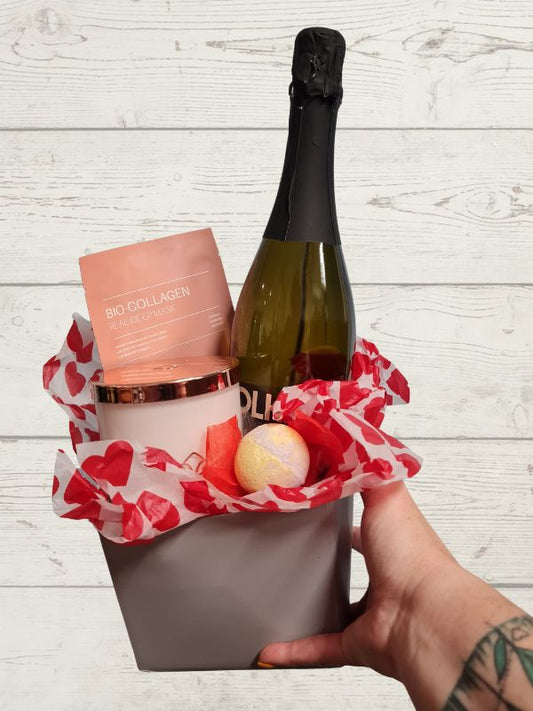 A gift box containing a bottle of non-alcoholic wine, a candle, a facemask, and a bath bomb, decorated with red and white paper with heart patterns.