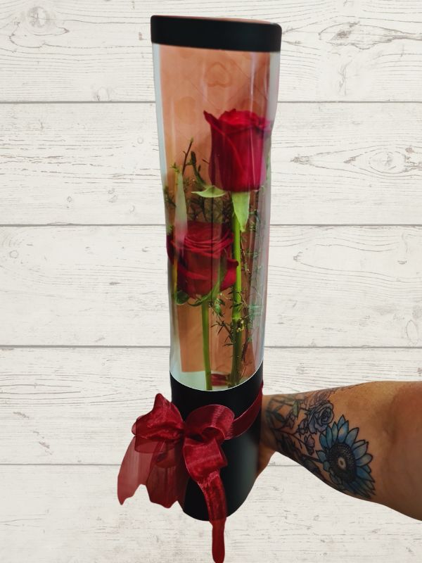 Two red roses inside a clear cylindrical vase with a red ribbon decoration, gift wrapped.