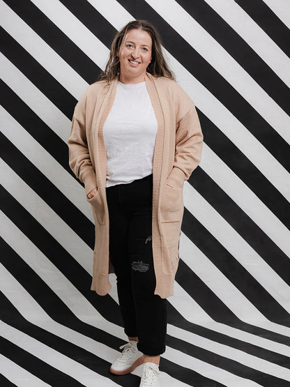 A woman standing against a black and white striped background, wearing a beige long knit open cardigan with bell sleeves, side splits, and ribbed edging, paired with a white t-shirt and black pants.