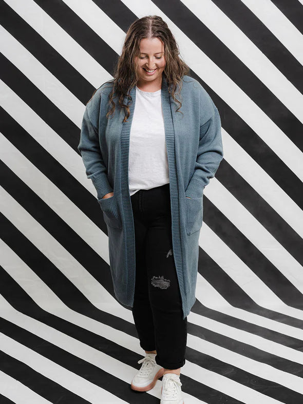 A woman standing against a black and white striped background, wearing a blue long knit open cardigan with bell sleeves, side splits, and ribbed edging, paired with a white t-shirt and black pants.