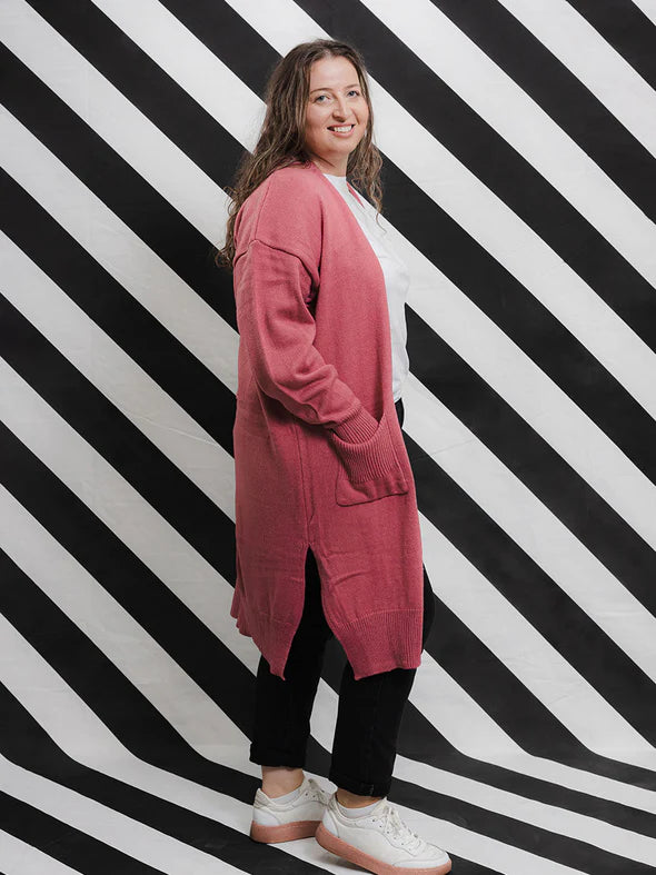 A woman standing against a black and white striped background, wearing a blush pink long knit open cardigan with bell sleeves, side splits, and ribbed edging, paired with a white t-shirt and black pants.
