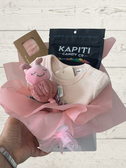 Baby gift set with pink plush toy, baby clothes, and Kapiti Candy Co packaging on a wooden surface.