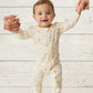 Baby in a cream onesie with small patterns standing on a light wooden floor.