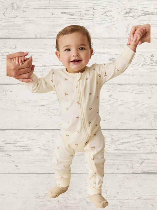 Baby in a cream onesie with small patterns standing on a light wooden floor.
