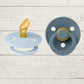 Two BIBS pacifiers with an anatomical glow design, one pacifier with a green ring on the shield and the other with a black ring, placed on a textured surface.