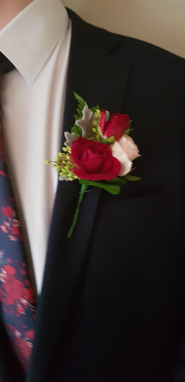 A close-up of a formal boutonniere flower bouquet with a red rose, pink flower, and green leaves pinned to a black suit jacket on a person wearing a white dress shirt.